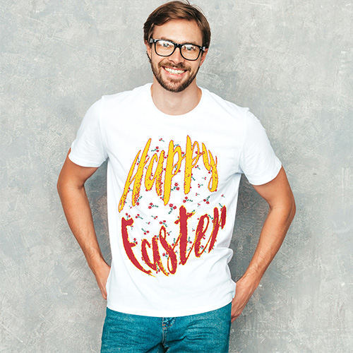 A smiling man wearing white T-shirt with red and yellow “Happy Easter” brush lettering, standing casually against a grey wall.