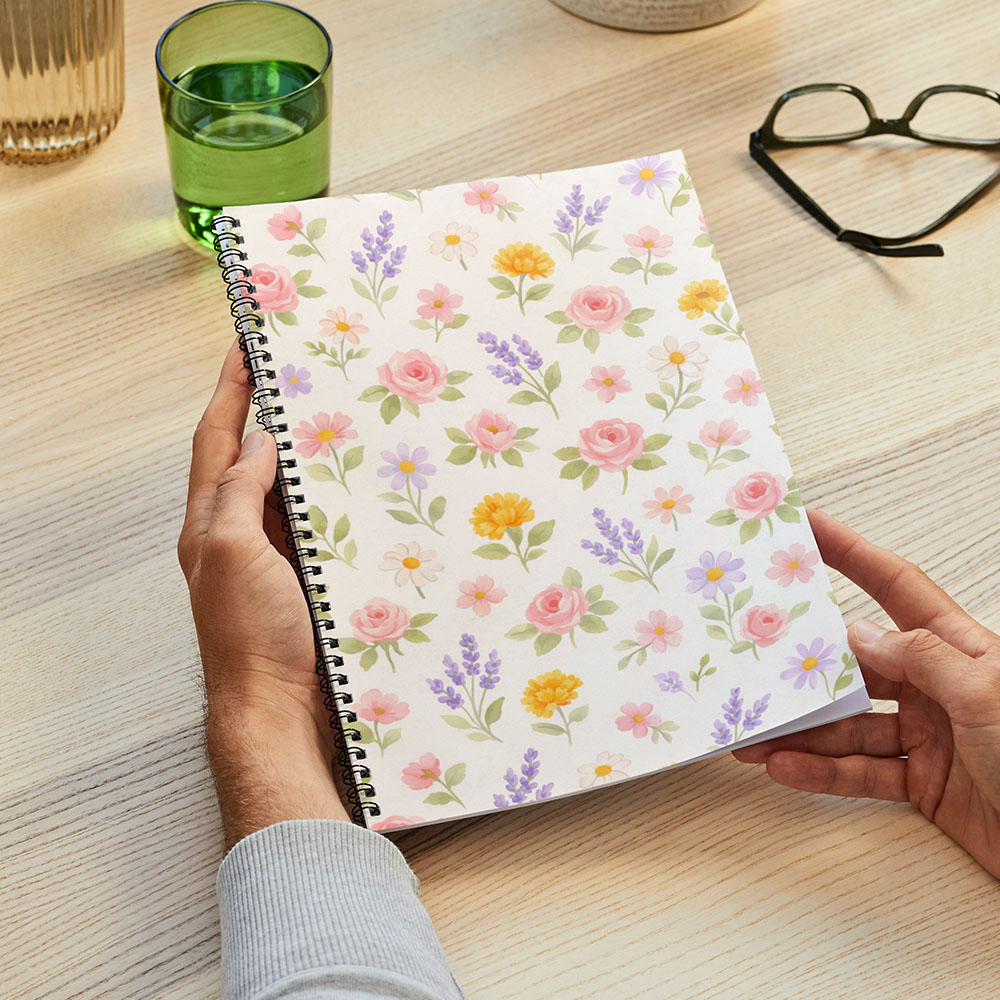 Hands holding a spiral notebook covered in a pastel watercolor floral pattern, styled on a desk for creative journaling use.